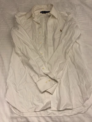 Vit skjorta från Ralph Lauren - Skjortan är i jätte bra skick behöver bara strykas. Klassisk vit skjorta från Ralph Lauren med broderad logga på bröstet. Skjortan har lång ärm, knappar framtill och är tillverkad i bomull. Perfekt till en clean och stilren look.
