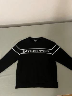 Svart EA7 Emporio Armani sweatshirt - Svart sweatshirt från EA7 Emporio Armani med vita detaljer och logga framtill. Tröjan har rund halsringning, långa ärmar och vita linjer över bröstet och axlarna. Perfekt för en stilren och sportig look.