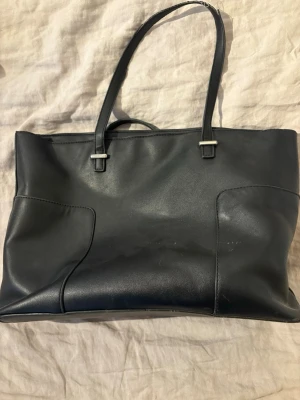 Svart axelväska i skinnimitation - Stilren svart axelväska med raka linjer och dubbla axelband. Väskan har en enkel design utan synliga loggor och är tillverkad i skinnimitation. Perfekt storlek för att få plats med laptop, böcker och annat viktigt. Många defekter men alla syns på bilderna.