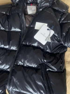 Svart glansig dunjacka från Moncler - Snygg svart dunjacka från Moncler med glansig finish och klassisk quiltad design. Jackan har dragkedja framtill, hög krage och en ikonisk Moncler-logga på insidan. Perfekt för kalla dagar och riktigt trendig look. Storlek 5