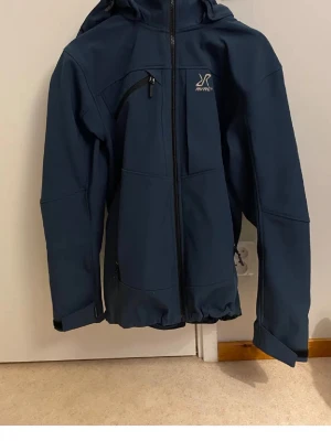 Mörkblå vindjacka från RYRC - Snygg mörkblå vindjacka från RYRC med huva och dragkedja framtill. Jackan har en bröstficka med dragkedja och diskret logga på bröstet. Perfekt för blåsiga dagar och enkel att matcha med olika outfits.