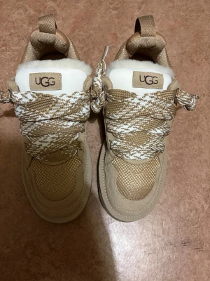 UGG LOWMEL - Snygga beige sneakers från UGG med grova, mönstrade snören i beige och vitt. Skorna har en rund tå, platt sula och är tillverkade i mocka och textil med vit fluffig insida. Perfekta för dig som gillar en stilren och bekväm look.