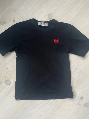 Svart Comme des Garçons PLAY t-shirt - Svart t-shirt från Comme des Garçons PLAY med det ikoniska röda hjärtat broderat på bröstet. Klassisk rund hals och korta ärmar. Tillverkad i mjuk bomull för en skön och avslappnad look. XS/S