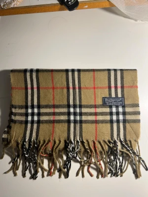 Beige rutig halsduk från Burberrys - Klassisk beige halsduk från Burberrys i 100% lammull med det ikoniska rutmönstret i svart, vitt och rött. Halsduken har fransar i ändarna och en snygg vävd etikett. Perfekt accessoar för att lyfta din outfit.