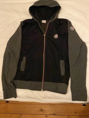 Svart och grå hoodie från Moncler - Snygg hoodie från Moncler i svart och grått med dragkedja framtill. Hoodien har huva, två fickor och Moncler-logga på bröstet och ärmen. Materialet är en mix av mesh och mjuk bomull, vilket ger en sportig och modern look. Perfekt för dig som gillar streetwear. Dock har dragkedjan lossnat men den funkar fortfarande lika bra och är enkel att fixa!