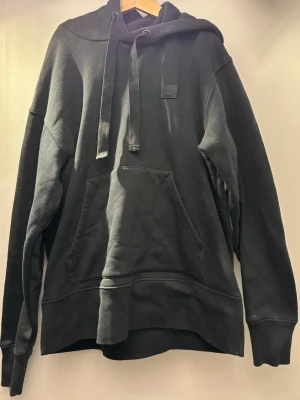 Svart hoodie med ficka och huva - En stilren svart hoodie från acne studios med stor ficka framtill och justerbar huva med breda snören. Hoodien har ribbade muddar vid ärmslut och nederkant samt en liten patch på bröstet. Perfekt för en avslappnad och cool look. Hoodien är använd ett fåtal gånger och är därför i ett mycket bra skick!