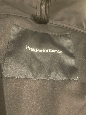 Svart vindjacka från Peak Performance - Snygg svart vindjacka från Peak Performance med  text design på vänstra armen. Fickorna går inte att zipa upp men går att fixa. Små skador på jackan.