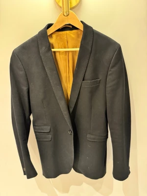 Svart kavaj från Vailent - Snygg svart kavaj från Vailent med klassisk passform och guldfärgat innerfoder. Kavajen har tre fickor fram, en knappstängning och långa ärmar. Perfekt för dig som vill ha en stilren och tidlös look.