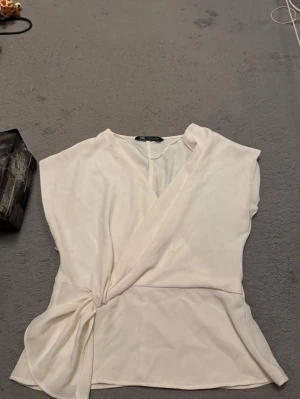 Vit omlottopp från Zara - Säljer en stilren vit omlottopp från Zara i mjukt, glansigt material. Toppen har ärmlös design, knyt i sidan och en snygg v-ringning framtill. Perfekt att styla med jeans eller kjol för en clean look.