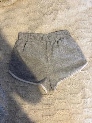 Grå mjuka shorts med resårmidja - Grå shorts i mjukt bomullsmaterial med elastisk midja och vita kanter i storlek XS (passar S) Klassisk sportig look med loose fit och korta ben, perfekta för sommardagar eller chill hemma. Inga fickor eller synliga loggor.