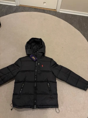 Svart pufferjacka från Polo Ralph Lauren - Svart pufferjacka från Polo Ralph Lauren med röd broderad logga på bröstet. Jackan har huva, dragkedja framtill och två snedställda fickor med dragkedja. Quiltad design och justerbara snören nedtill för extra passform.