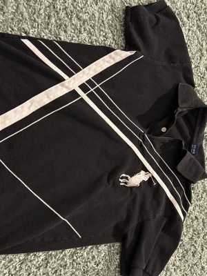 Svart pikétröja från Ralph Lauren - Svart pikétröja från Ralph Lauren med vita diagonala ränder och stor broderad logga på bröstet. Klassisk krage och korta ärmar. Tillverkad i bomull för en bekväm känsla och stilren look.