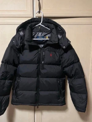 Svart pufferjacka från Polo Ralph Lauren - Säljer en svart pufferjacka från Polo Ralph Lauren med huva och dragkedja. Jackan har en röd broderad logga på bröstet, flera fickor med dragkedja och en riktigt fet, vadderad look. Perfekt för kalla dagar när du vill hålla stilen.