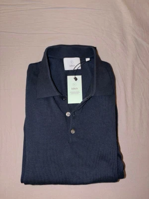 Mörkblå pikétröja från Bläck - Snygg mörkblå pikétröja från Bläck med klassisk krage och två knappar framtill. Tröjan är tillverkad i ett mjukt stickat material som ger en stilren och avslappnad look. Perfekt till jeans eller chinos för en clean outfit.