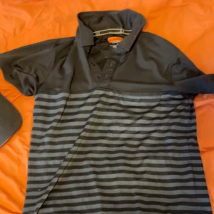 Svart och grå randig piké  - Säljer en svart och grå randig polotopp från Dockers. Toppen har krage, knappar framtill och korta ärmar. Perfekt för dig som gillar en enkel men stilren look med diskreta ränder.