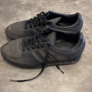 Svarta Adidas sneakers med mesh - Säljer ett par svarta Adidas sneakers med klassiska tre ränder på sidan och meshmaterial för bra ventilation. Skorna har snörning och platt sula, samt Adidas-logga på hälen. Perfekta för en sportig och avslappnad stil.