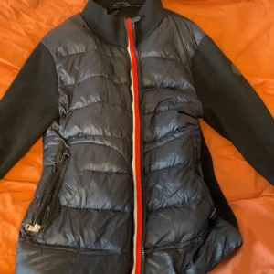 Svart teddyjacka från Moncler - Snygg svart teddyjacka från Moncler med quiltad front och stickade ärmar. Jackan har dragkedja med röd och vit kant, två fickor med dragkedja och Moncler-logga på ärmen. Perfekt för dig som vill ha en sportig och trendig look.