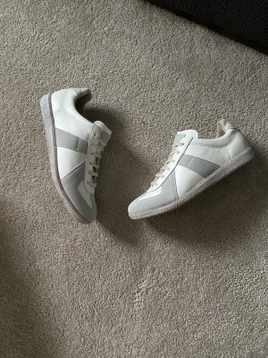 Maison Margiela White Paint Gats - Snygga Margielas i vitt och ljusgrått med klassisk design. Skorna har detaljer i mocka på sidan och framtill, samt snörning och platt sula. Perfekta för dig som gillar stilrena och tidlösa sneakers. Innersula på 30,5cm vilket är ungefär 46/47.