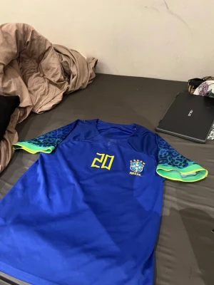 Blå Brasilien fotbollströja #20 - Snygg blå Brasilien fotbollströja med nummer 20 i gult på bröstet och CBF-emblemet broderat. Ärmarna har blått leopardmönster med gröna och gula detaljer. Perfekt för match eller träning, tillverkad i lätt och ventilerande material.