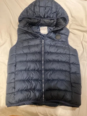 Mörkblå dunväst med huva Minoti - Snygg mörkblå dunväst från Minoti med vadderad design och praktisk huva. Västen har dragkedja framtill och en rund logotyp på bröstet. Perfekt för lager-på-lager och håller dig varm under kyliga dagar.