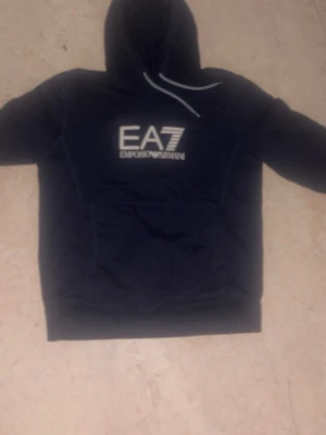 Svart EA7 Emporio Armani hoodie - Snygg blå hoodie från EA7 Emporio Armani med stor vit logga på bröstet. Klassisk huva med dragsko och magficka framtill. Perfekt för dig som gillar streetwear och vill ha en stilren look. Mjuk och bekväm bomullsblandning.