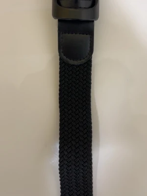 Svart flätat bälte med metallspänne - Snyggt svart bälte med flätad design och matchande svart metallspän sop. Bältet har detaljer i konstläder vid spännet och är flexibelt tack vare det elastiska materialet. Perfekt för att lyfta vilken outfit som helst.