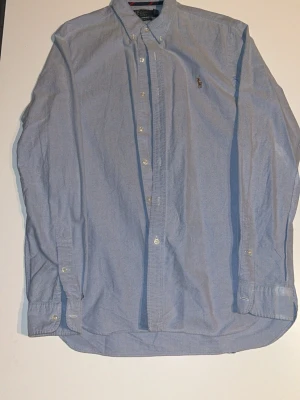 Blå skjorta från Polo Ralph Lauren M - Klassisk ljusblå skjorta från Polo Ralph Lauren i slim fit med broderad logga på bröstet. Skjortan har knappar hela vägen, button down-krage och långa ärmar. Tillverkad i bomull och passar perfekt till en clean och stilren look. den har lite defekter som är lagade.