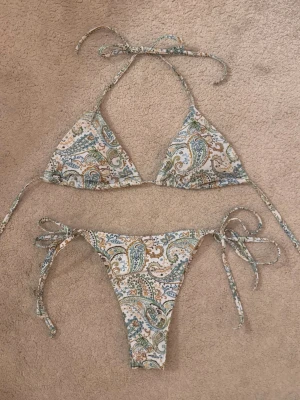 Paisleymönstrad trekantsbikini - Snygg bikini med paisleymönster i grönt, blått och gult på vit botten. Överdelen har trekantskupor med knyt i nacke och rygg, och underdelen har knyt i sidorna för justerbar passform