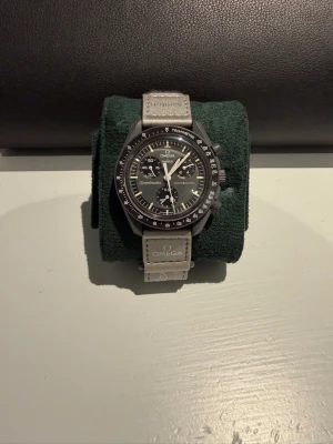 Omega Speedmaster svart kronograf - Snygg Omega Speedmaster kronograf med svart urtavla och vit/silverfärgade detaljer. Klockan har tachymeter-skala på bezeln och grått läderarmband med Omega-logga. Klassisk rund form och sportig design, perfekt för dig som gillar stilrena accessoarer.