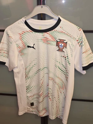 Portugal fotbollströja Puma vit - Säljer en vit Portugal fotbollströja från Puma med svart krage och ärmslut. Tröjan har ett färgglatt mönster i grönt, rött och orange samt Portugals landslagsemblem på bröstet. Perfekt för dig som älskar fotboll och vill stötta Portugal på match! Den är använd 1 gång och den är tvättad och är ute för försäljning. Hör av er om ni har frågor! Priset är ej hugget i sten. 