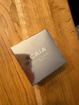CAIA Setting Powder Glow puder - Säljer ett CAIA Setting Powder Glow i en fyrkantig, spegelblank förpackning. Pudret har en varm beige ton och ger en glowig finish. Perfekt för att fixera foundation och ge huden extra lyster. Kommer i en praktisk dosa med sil för enkel applicering. Endast testad!
