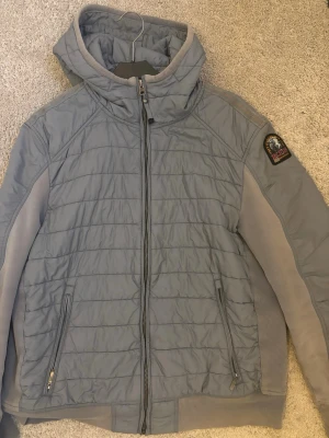  Dunjacka från Parajumpers - Snygg grå dunjacka från Parajumpers med huva och quiltad design. Jackan har dragkedja framtill, två sidofickor med dragkedja och en patch på ärmen. Perfekt för kalla dagar och har en sportig vibe. Är storlek L men passar M 