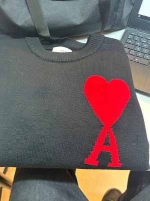 Svart stickad tröja från Ami Paris - Säljer en svart stickad tröja från Ami Paris med deras ikoniska röda hjärta och bokstaven A på bröstet. Tröjan har rund halsringning och ribbade muddar. Perfekt för dig som gillar stilrena plagg med en twist. Bara använt 2 gånger.