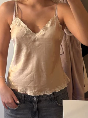 Champagnefärgad linnetopp med spets - Snygg champagnefärgad linnetopp med tunna axelband och v-ringning. Toppen har fina spetsdetaljer vid halsringning och nederkant. Jag är 180cm lång och bär vanligtvis S/M. Skicka gärna frågor eller prisförslag!