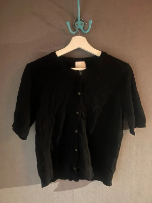 Svart kortärmad kofta - Stilren svart kofta från H&M med korta ärmar och rund halsringning. Koftan har knäppning framtill med svarta knappar och är tillverkad i ett mjukt, tunt material som passar perfekt för lager på lager. Enkel och klassisk design som funkar till mycket. Lite katthår på sig. Står m men passar s också. Använd 2 gånger.
