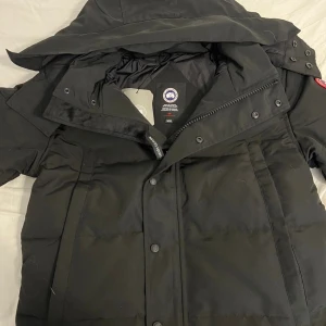 Svart dunjacka från Canada Goose - Säljer en svart dunjacka från Canada Goose med huva och klassisk röd logotyp på ärmen. Jackan har dragkedja och knappar framtill, samt flera praktiska fickor. Perfekt för kalla dagar med sin tjocka och puffiga design. Stilsäker look och grym komfort.