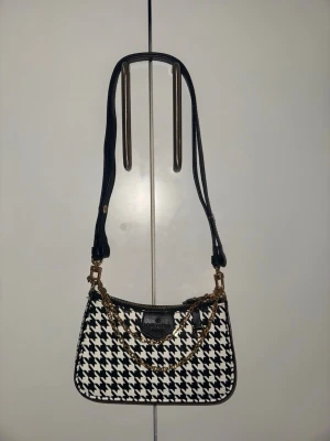  svartvit axelväska - Säljer en snygg axelväska i svartvitt hundtandsmönster. Väskan har guldfärgade kedjedetaljer, justerbart svart axelband och dragkedja upptill. Märkeslogga framtill och stilren form som ger en trendig vibe.