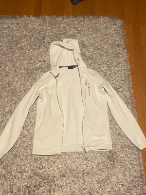 Vit hoodie från Sail Racing - Vit hoodie från Sail Racing med dragkedja framtill och en praktisk bröstficka med dragkedja. Hoodien har broderad logga på bröstet och är tillverkad i ett mjukt fleecematerial. Perfekt för en sportig och avslappnad stil.