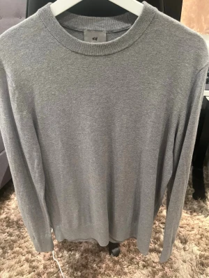Grå stickad kashmir tröja från H&M - En stilren grå stickad kashmir tröja från H&M med rund halsringning och långa ärmar. Tröjan har ribbade muddar vid hals, ärmslut och nederkant. Perfekt basplagg för lager på lager och enkel att matcha med olika outfits.