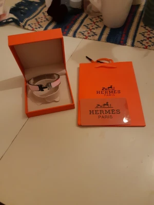 Rosa HERMÈS Clic H armband - Säljer ett ikoniskt HERMÈS Clic H armband i ljusrosa emalj med silverfärgad metall och klassiskt H-spänne. Kommer i original ask och med HERMÈS påse och kort. Perfekt accessoar för dig som gillar lyxiga detaljer och stilren design.