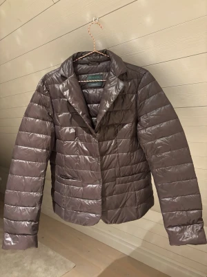 Brun dunväst från Benetton - Snygg brun dunväst från Benetton med glansig finish och klassisk krage. Västen har horisontella sömmar, två fickor framtill och stängs med knappar. Perfekt och ger en stilren look till hösten och vintern.