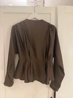 Brun peplumblus med lång ärm - Snygg brun blus med peplumskärning och långa ärmar. Blusen har en markerad midja och dragkedja bak för enkel påklädning. Tillverkad i ett mjukt, lätt tyg som ger ett fint fall.