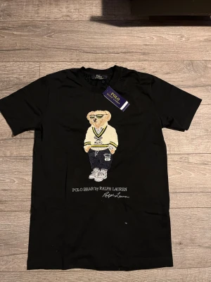 Polo bear by Ralph Lauren  - Polo Ralph Lauren T-shirt med ikoniska Polo Bear-motivet. Svart, klassisk passform i mjuk bomull. Fick den som gåva från min far när han var i Frankrike 🇫🇷 , aldrig använd – originaltag kvar.  Perfekt nu till sommaren 🔥