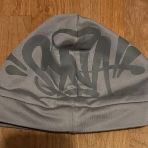 Grå Syna World mössa - Säljer en grå Syna World mössa med ett stort, grafiskt tryck på framsidan. Materialet känns mjukt och följsamt, perfekt för att addera en cool touch till din outfit. Kan pratas om priset.