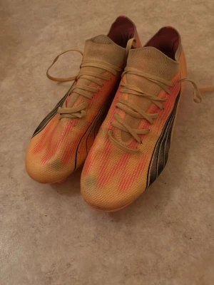 Puma fotbollskor - Säljer ett par riktigt snygga fotbollsskor i orange och rosa med svarta detaljer. Skorna har snörning och en ovandel i syntetmaterial som formar sig efter foten. Sulan är rosa med fasta dobbar för bra grepp på planen. Perfekta för dig som vill sticka ut på matchen!
