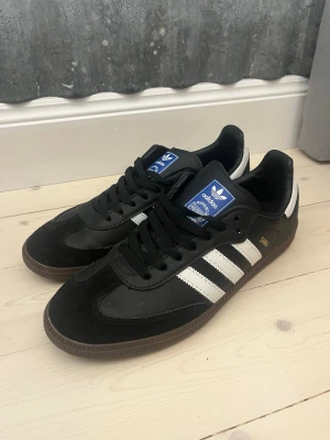Adidas Samba svart/vit sneakers - Klassiska Adidas Samba sneakers i svart med vita ränder och brun gummisula. Skorna har snörning, rund tå och detaljer i mocka och skinn. Perfekta för dig som gillar retrostil och streetwear. Adidas-logga på plösen och guldfärgad Samba-text på sidan.