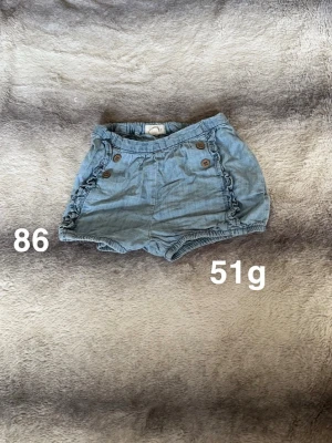 Blå shorts med volang och knappar - Säljer ett par ljusblå shorts med volangdetaljer längs sidorna och dekorativa bruna knappar framtill. Shortsen har resår i midjan och bensluten för en bekväm passform. Perfekta för varma dagar!🌸 Fint använt skick. Storlek 86