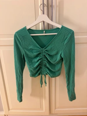 Grön ribbad topp från ONLY - Säljer en grön croppad topp från ONLY med långa ärmar och v-ringning. Toppen har ribbad struktur och snygg snörning framtill som ger en rynkad effekt. Perfekt till jeans eller kjol för en trendig look.