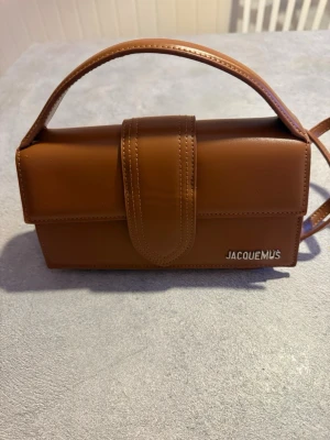 Brun axelväska från Jacquemus - Säljer en snygg brun axelväska från Jacquemus i slätt skinn. Väskan har ett stilrent, rektangulärt format med ett brett lock och justerbar axelrem. Metallogo framtill och insida i beige tyg. Perfekt för dig som vill ha en trendig och clean look. Nypris 10500 men priset jag säljer den för är pågrund av sminkfläckarna och hår från Katt i väskan. 