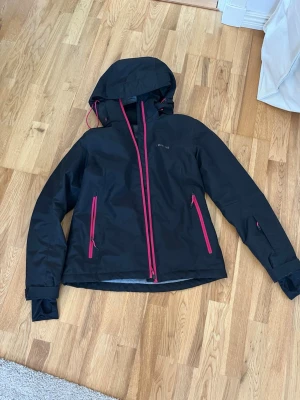 Svart vindjacka från Everest, strl 36 - Säljer en svart vindjacka från Everest i storlek 36. Jackan har huva, långa ärmar och rosa dragkedjor som snygg kontrast. Perfekt för blåsiga dagar och outdoor-äventyr. Materialet är slitstarkt och vattenavvisande syntet.
